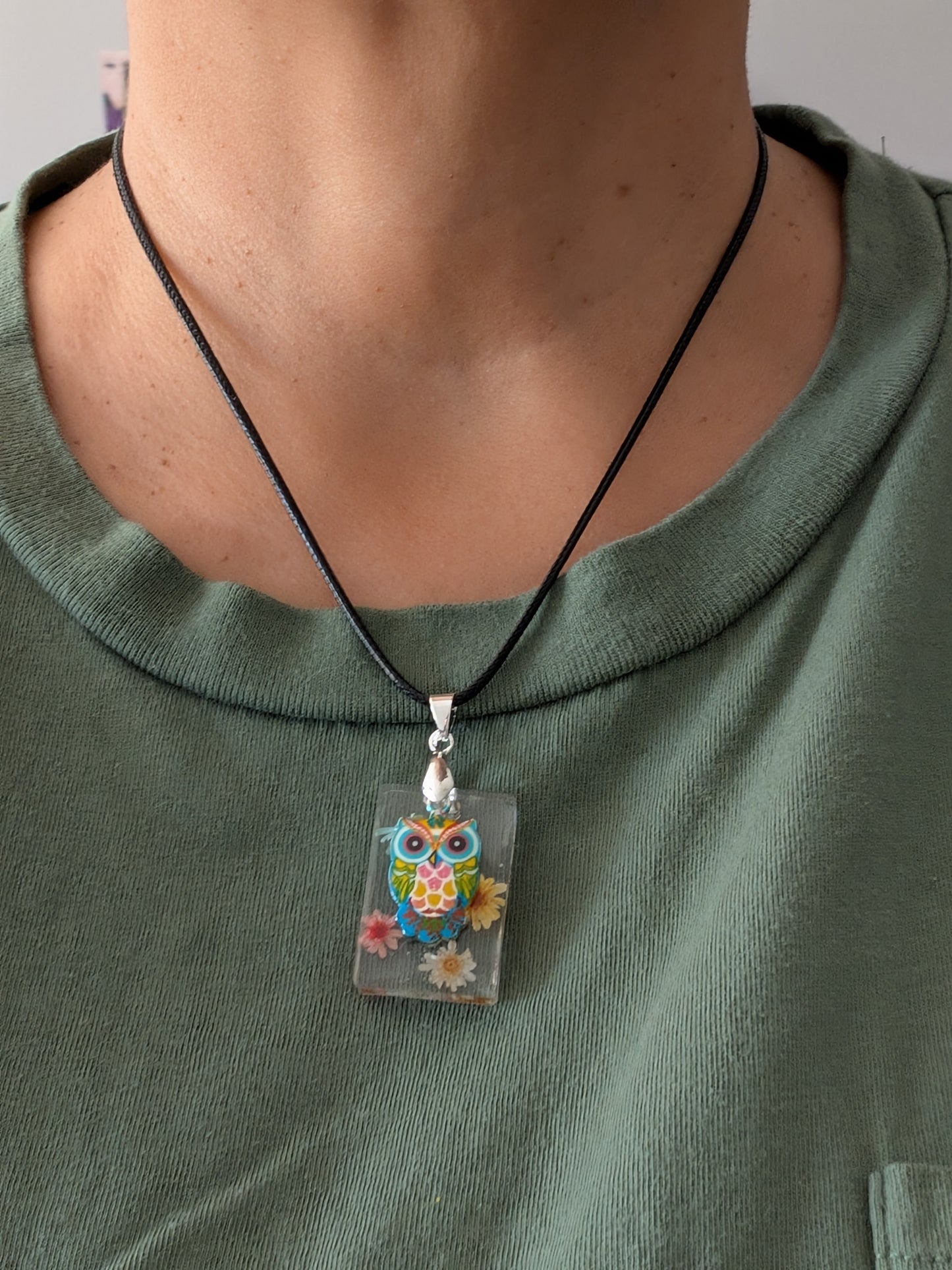 This 17.5-inch necklace features a unique rectangular resin pendant embedded with delicate flowers, paired with a charming silver owl accent. Suspended on a sleek black leather cord with a secure clasp, it combines natural beauty and subtle elegance, making it a perfect accessory to enhance any outfit.