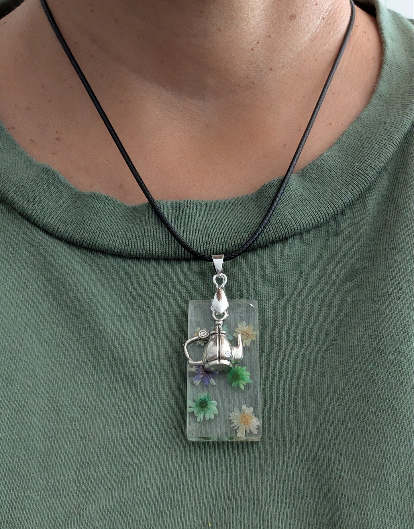 This 17.5-inch necklace features a unique rectangular resin pendant embedded with delicate flowers, paired with a charming silver kettle accent. Suspended on a sleek black leather cord with a secure clasp, it offers a stylish blend of nature-inspired beauty and modern design, perfect for adding a distinctive touch to any outfit.