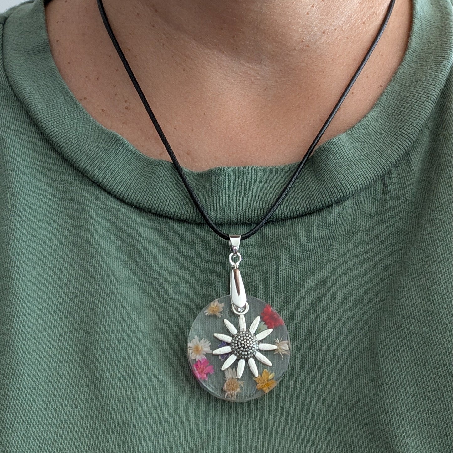 This elegant 17.5-inch necklace features a delicate resin circle embedded with vibrant flowers, complemented by a silver flower charm. Suspended on a sleek black leather cord with a secure clasp, it offers a perfect blend of natural beauty and modern style. Ideal for adding a subtle yet captivating touch to any outfit.