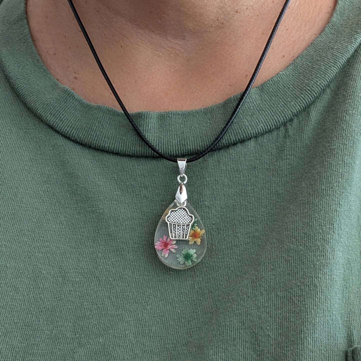 This charming 17.5-inch necklace showcases a resin teardrop filled with delicate flowers, paired with a playful silver cupcake charm. Set on a black leather cord with a secure clasp, it combines whimsical detail with stylish simplicity, making it a unique accessory to brighten any look.