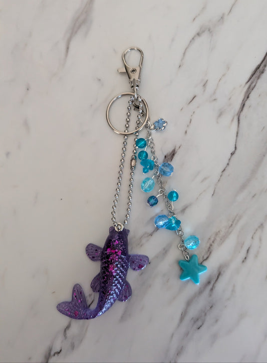 Decorative keychain/bag charm with purple fish charm and blue beads on a marble surface