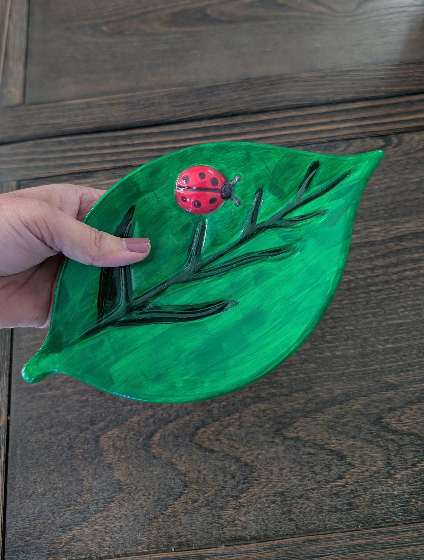 Hand holding a green leaf-shaped object with a red ladybug on a wooden surface