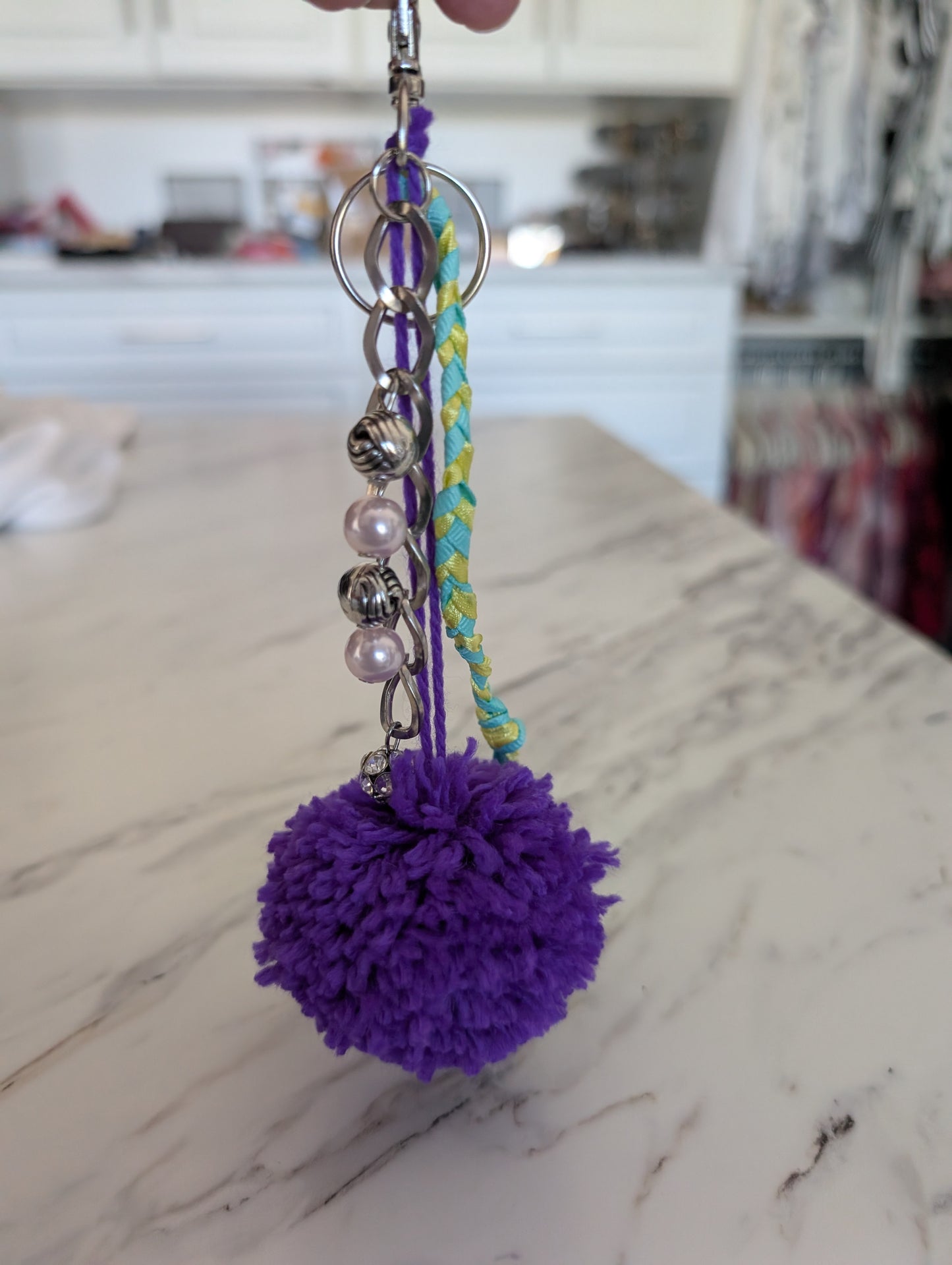 Purple pom-pom keychain with beads and a braided string on a marble surface.