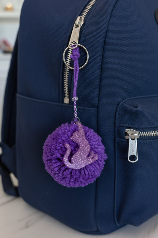 Purple pom-pom keychain with a cat design attached to a blue backpack.