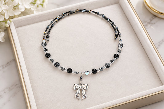 A beaded necklace with black and silver beads and a silver bow charm, displayed on a cream-colored jewelry box.