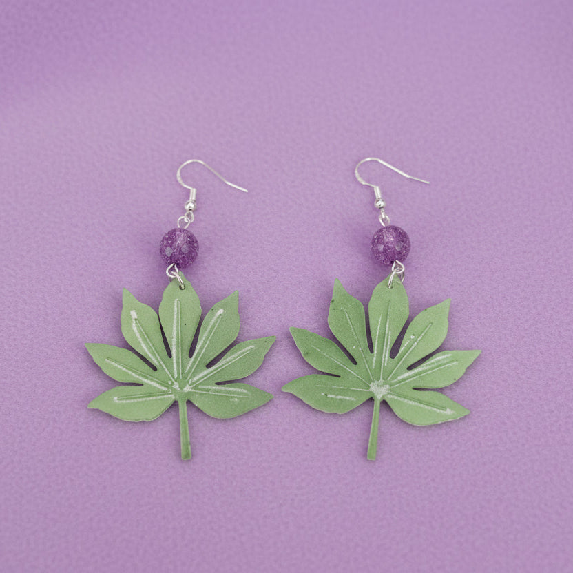 A pair of handmade dangle earrings featuring green leaf-shaped charms with white glitter accents, suspended from silver fishhook wires with a small purple glitter bead in between.