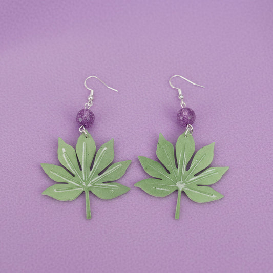 A pair of handmade dangle earrings featuring green leaf-shaped charms with white glitter accents, suspended from silver fishhook wires with a small purple glitter bead in between.