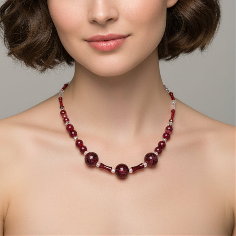 A close-up of a woman wearing a beaded necklace with deep red round beads, faceted red center beads, and small clear crystal accents against a neutral gray background.