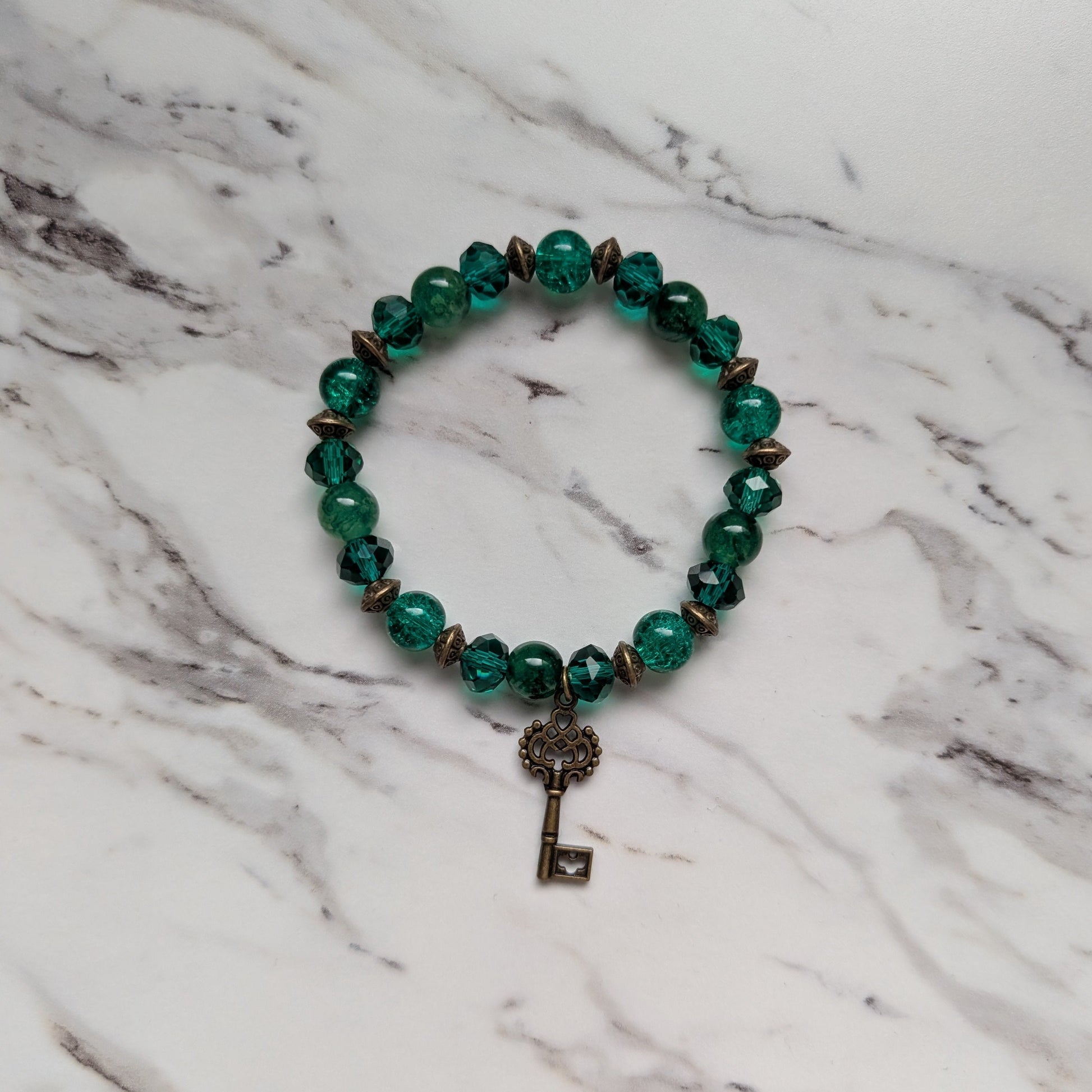 A beaded stretch bracelet featuring faceted green beads, small metallic bronze cone spacers, and a dangling vintage-style key charm, displayed on a white and gray marble surface.