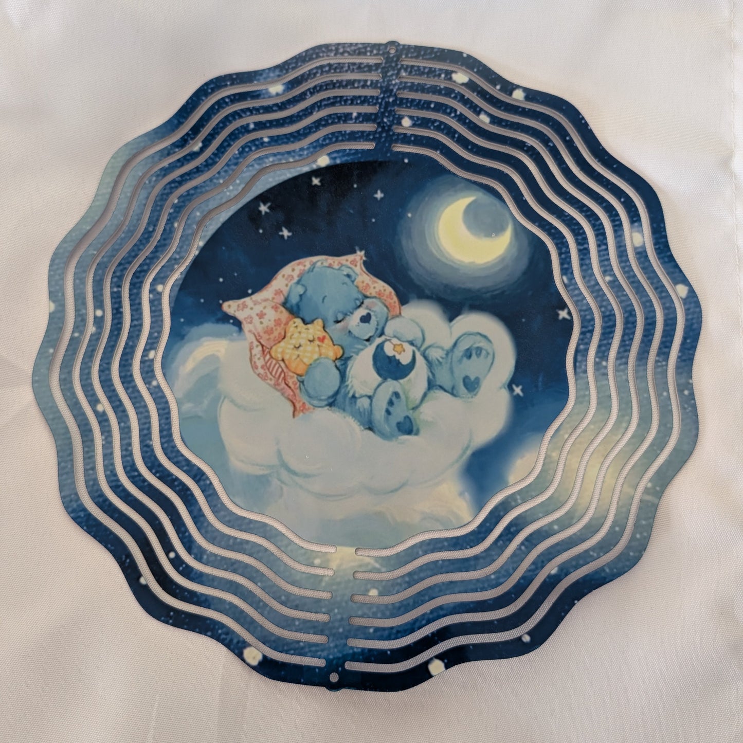 Bring whimsical charm to your outdoor space with this delightful 10-inch wind spinner showcasing Bedtime Care Bear on both sides. As it spins gracefully, stunning 4-inch teardrop crystal prisms dance with light, casting rainbow sparkles across your garden. Crafted from lightweight yet robust aluminum with a protective powder-coated finish, this spinner laughs in the face of rust and corrosion. The UV-treated image stays vibrant and cheerful through countless sunny days. Safe, smooth edges and hassle-free se