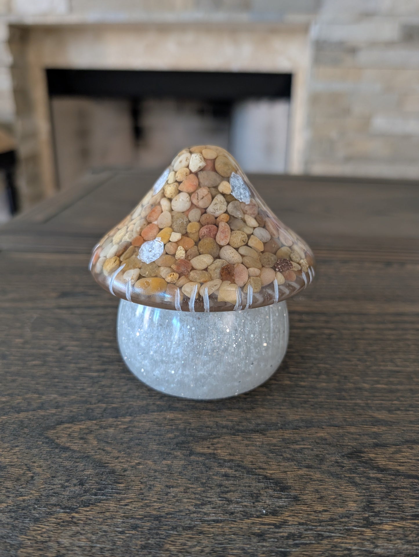 Decorative mushroom with pebbles on top on a wooden surface with a stone fireplace in the background.