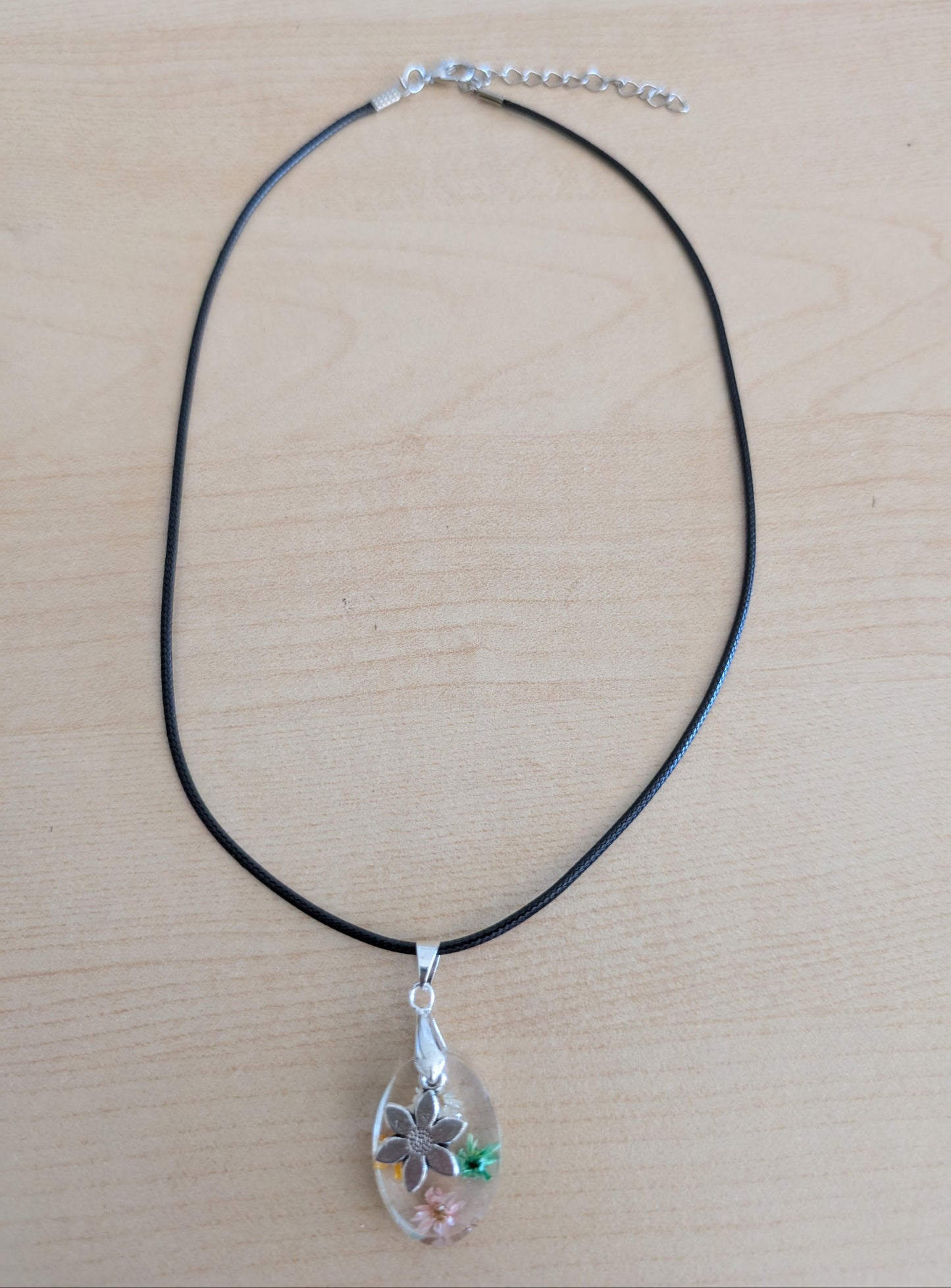 This 17.5-inch necklace showcases a stunning oval resin pendant filled with delicate flowers, complemented by a silver flower charm. Strung on a black leather cord with a secure clasp, it offers a unique blend of natural elegance and modern style, perfect for enhancing any outfit with a touch of floral beauty.