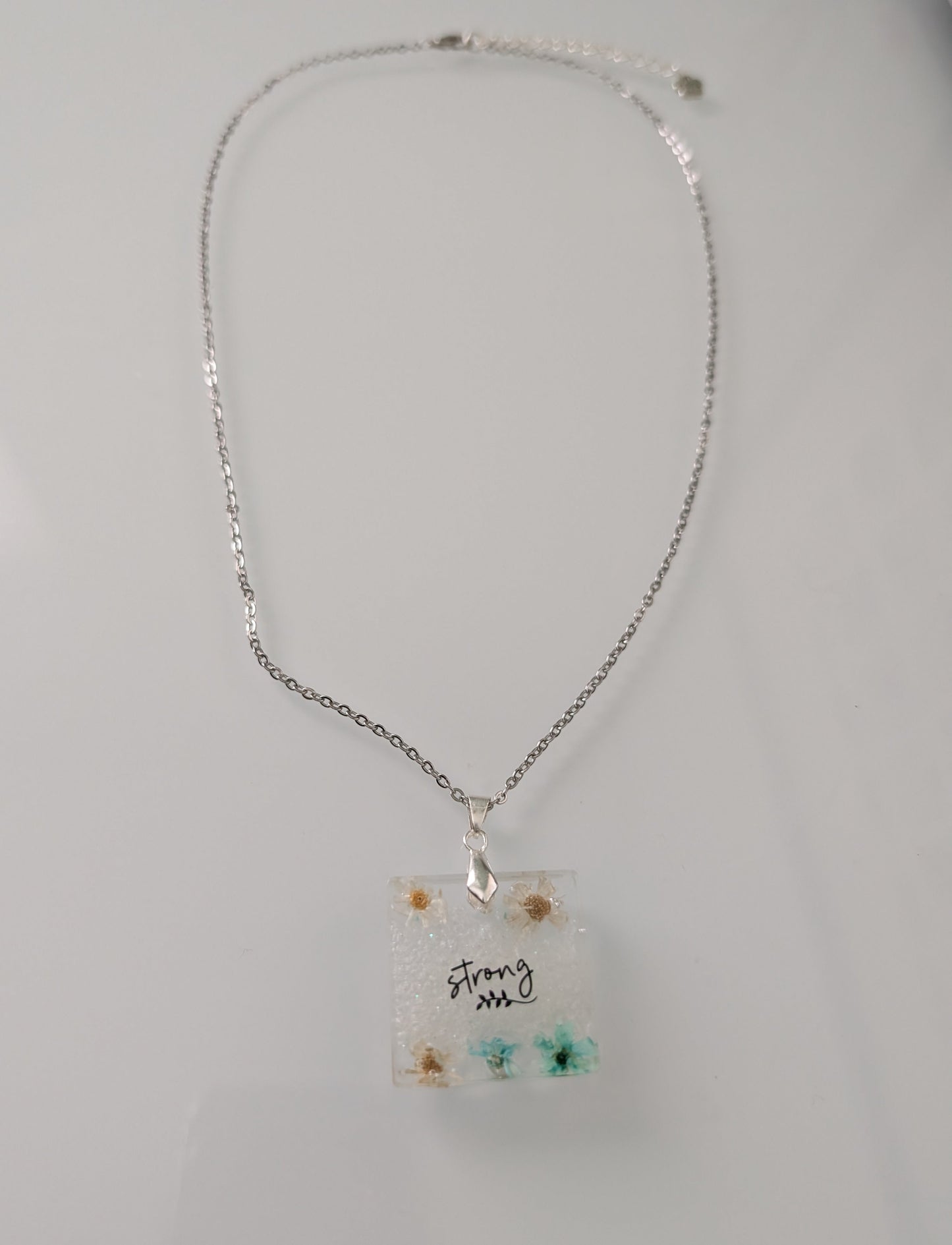This stainless steel cable link chain necklace features a beautiful square pendant with real flowers embedded in resin. The necklace is 21 inches long, with an additional 2 inch extension chain. Make a statement with this one-of-a-kind piece!

Necklace - 21 inches stainless steel cable link chain with a resin with flowers square pendant