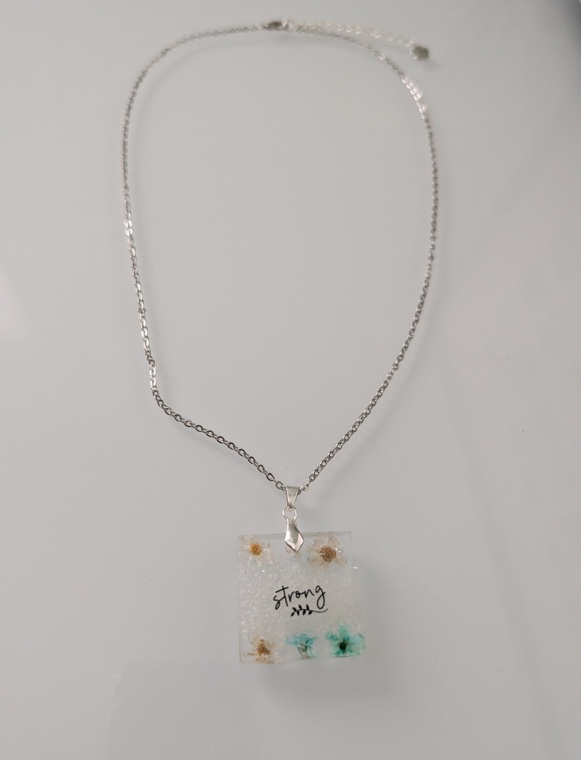 This stainless steel cable link chain necklace features a beautiful square pendant with real flowers embedded in resin. The necklace is 21 inches long, with an additional 2 inch extension chain. Make a statement with this one-of-a-kind piece!

Necklace - 21 inches stainless steel cable link chain with a resin with flowers square pendant