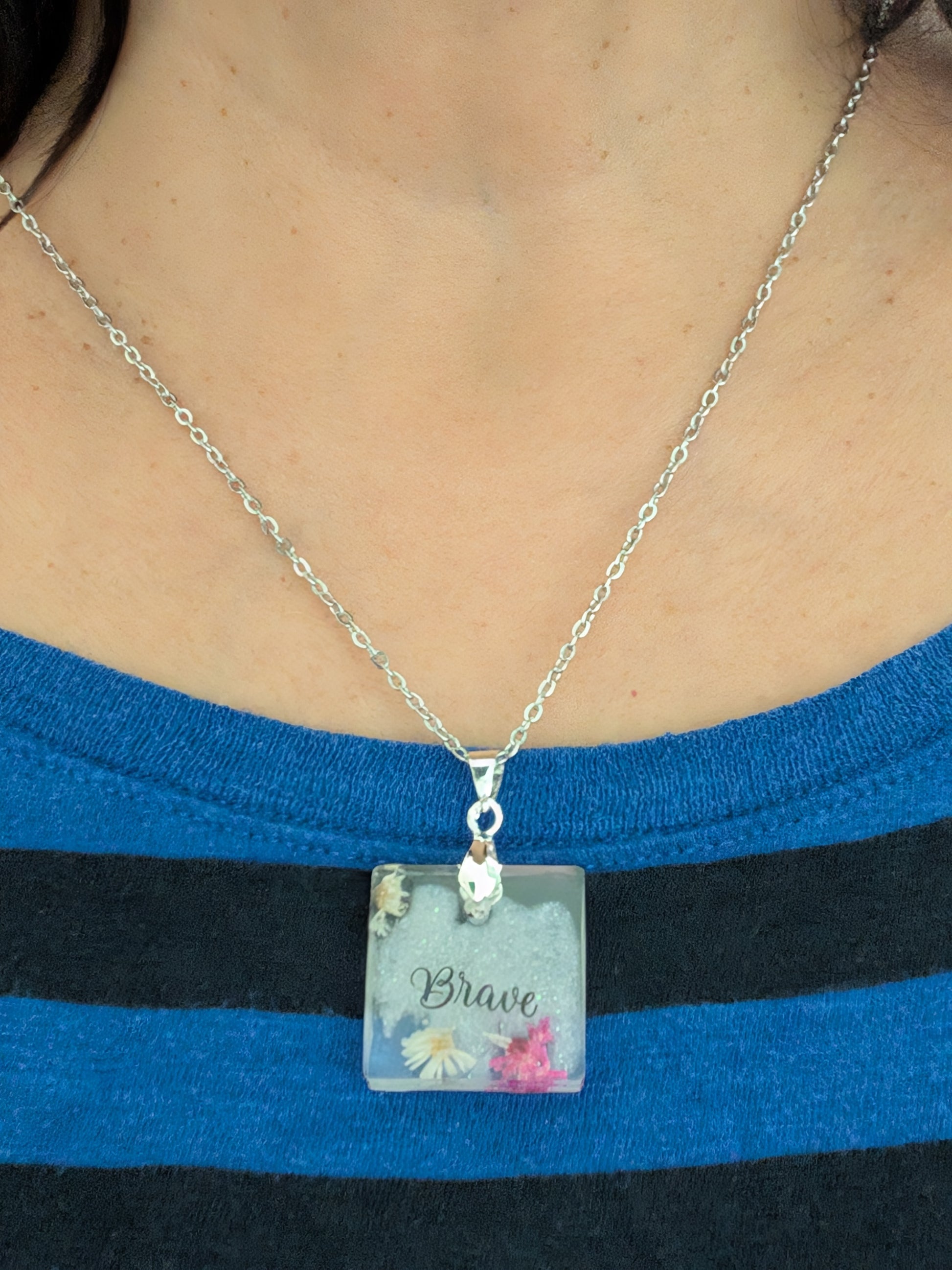 This stainless steel cable link chain necklace features a beautiful square pendant with real flowers embedded in resin. The necklace is 21 inches long, with an additional 2 inch extension chain. Make a statement with this one-of-a-kind piece!

Necklace - 21 inches stainless steel cable link chain with a resin with flowers square pendant