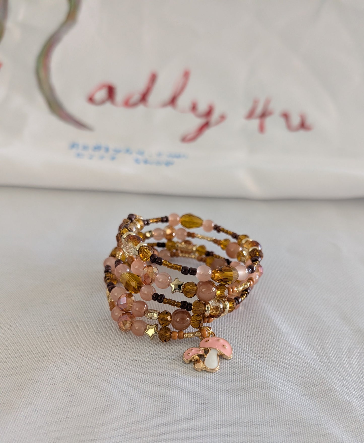 Elevate your wrist with this charming memory wire bracelet featuring a delightful blend of earthy tones and whimsical details. The four-coil design showcases beautiful 6mm tan and pink glass and crystal beads paired with warm gold, tan, and brown spacer accents. A sweet pink mushroom charm adds a playful touch that makes this piece perfect for nature lovers and charm collectors alike. Memory wire ensures a comfortable, adjustable fit that works for most wrist sizes—no clasp needed. Whether you're dressing u