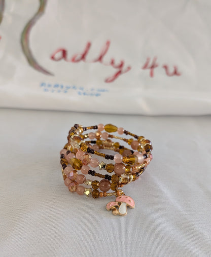Elevate your wrist with this charming memory wire bracelet featuring a delightful blend of earthy tones and whimsical details. The four-coil design showcases beautiful 6mm tan and pink glass and crystal beads paired with warm gold, tan, and brown spacer accents. A sweet pink mushroom charm adds a playful touch that makes this piece perfect for nature lovers and charm collectors alike. Memory wire ensures a comfortable, adjustable fit that works for most wrist sizes—no clasp needed. Whether you're dressing u