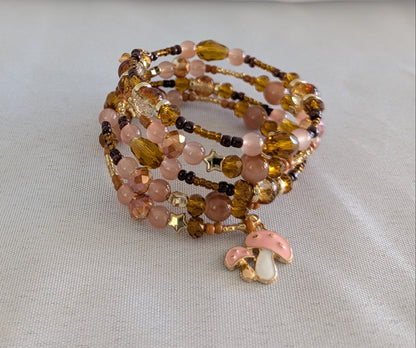 Elevate your wrist with this charming memory wire bracelet featuring a delightful blend of earthy tones and whimsical details. The four-coil design showcases beautiful 6mm tan and pink glass and crystal beads paired with warm gold, tan, and brown spacer accents. A sweet pink mushroom charm adds a playful touch that makes this piece perfect for nature lovers and charm collectors alike. Memory wire ensures a comfortable, adjustable fit that works for most wrist sizes—no clasp needed. Whether you're dressing u