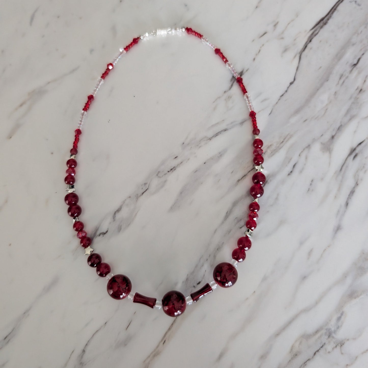 A beaded necklace with deep red faceted beads and clear spacer beads, laid flat on a white and gray marble surface.