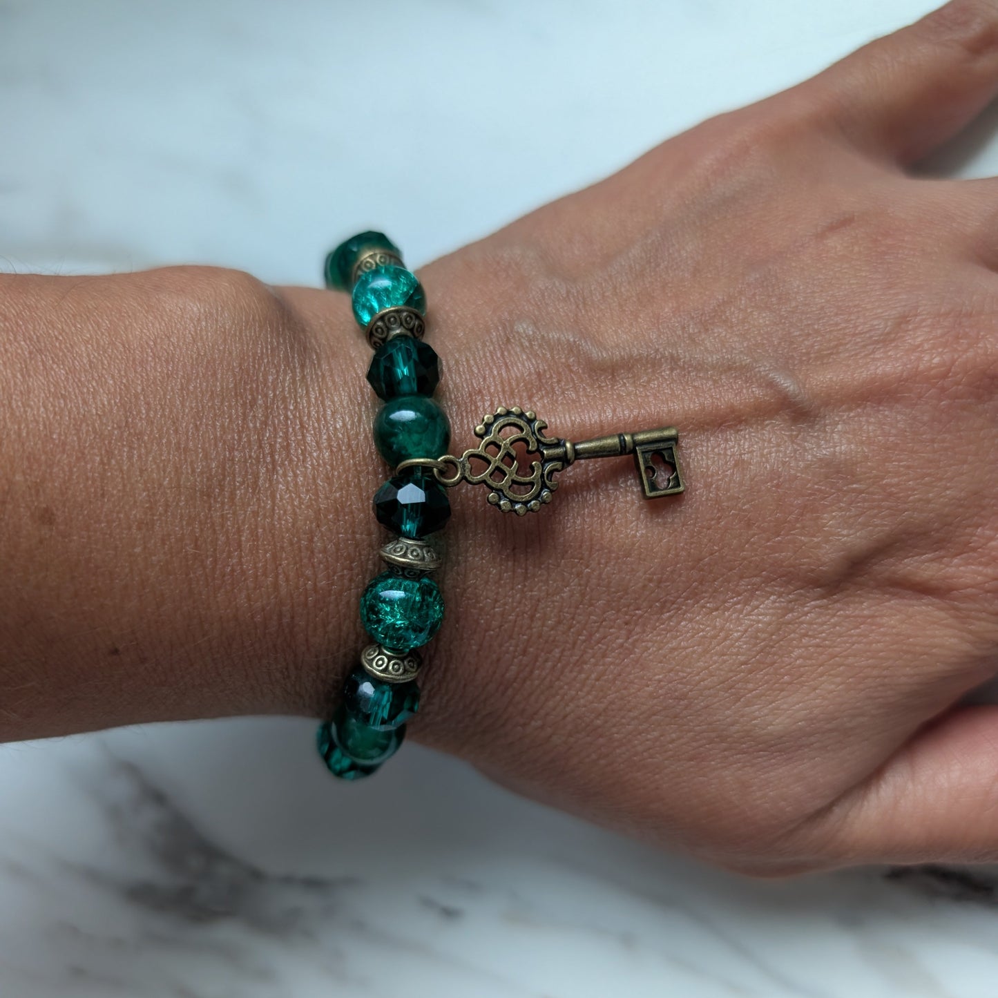 A close-up of a person's wrist wearing a beaded bracelet with dark green faceted beads, small bronze spacer accents, and a dangling antique-style key charm.