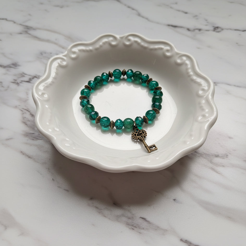 A beaded stretch bracelet with translucent green beads and a small antique-style key charm, displayed on a white scalloped ceramic dish over a white marble surface.