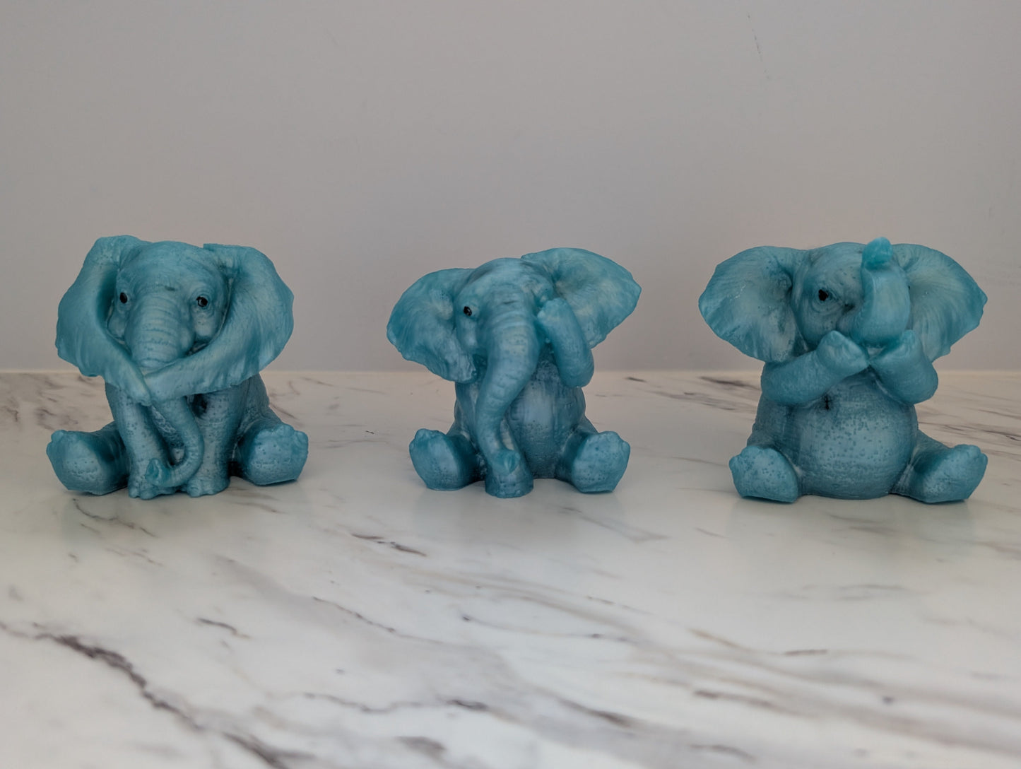 Handmade set of three blue resin baby elephant figurines representing see no evil, hear no evil, and say no evil on a marble surface.