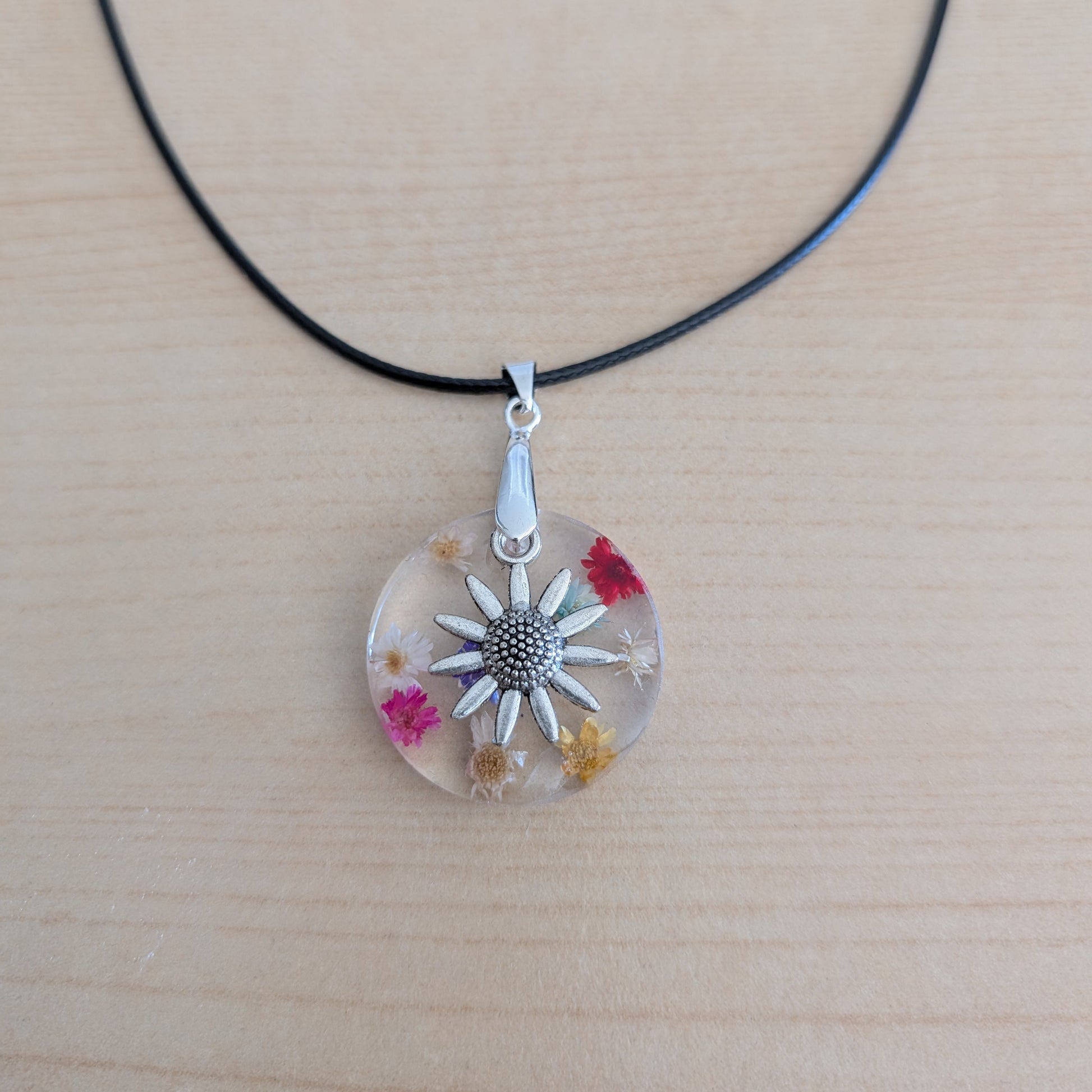 This elegant 17.5-inch necklace features a delicate resin circle embedded with vibrant flowers, complemented by a silver flower charm. Suspended on a sleek black leather cord with a secure clasp, it offers a perfect blend of natural beauty and modern style. Ideal for adding a subtle yet captivating touch to any outfit.