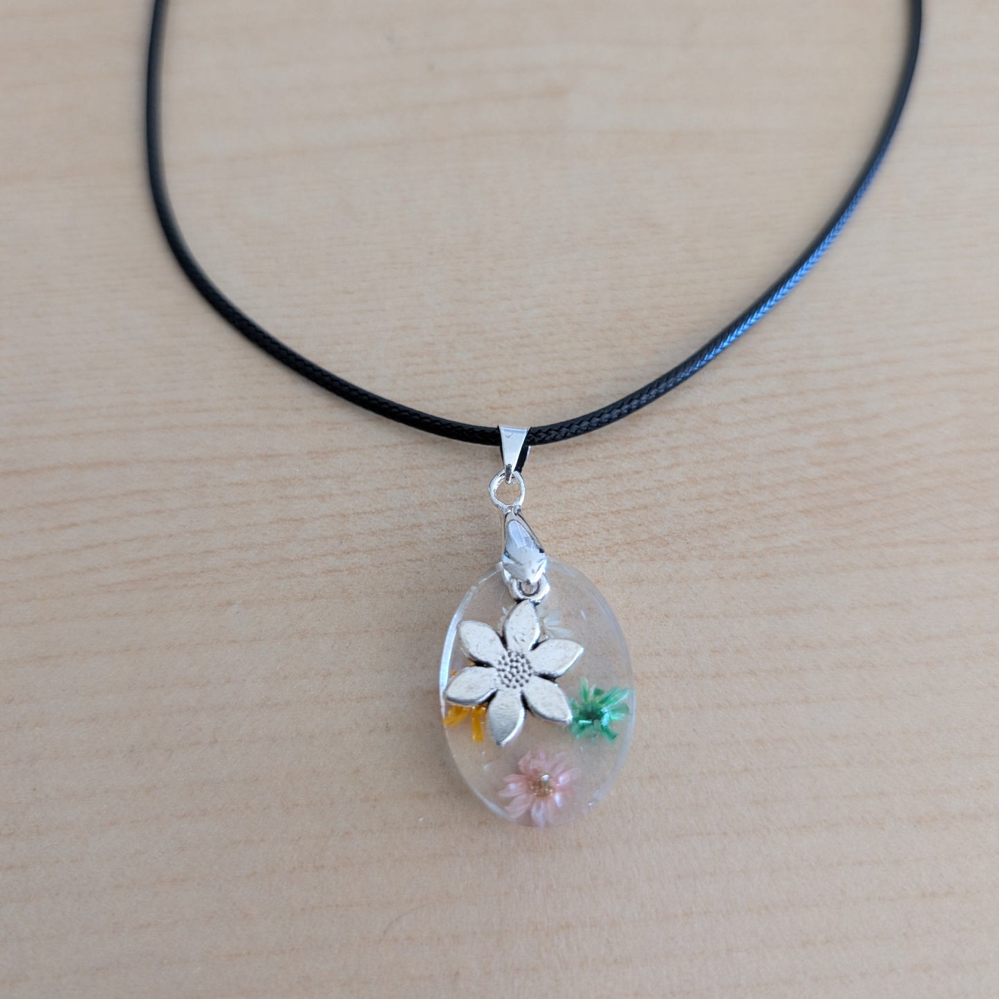 This 17.5-inch necklace showcases a stunning oval resin pendant filled with delicate flowers, complemented by a silver flower charm. Strung on a black leather cord with a secure clasp, it offers a unique blend of natural elegance and modern style, perfect for enhancing any outfit with a touch of floral beauty.