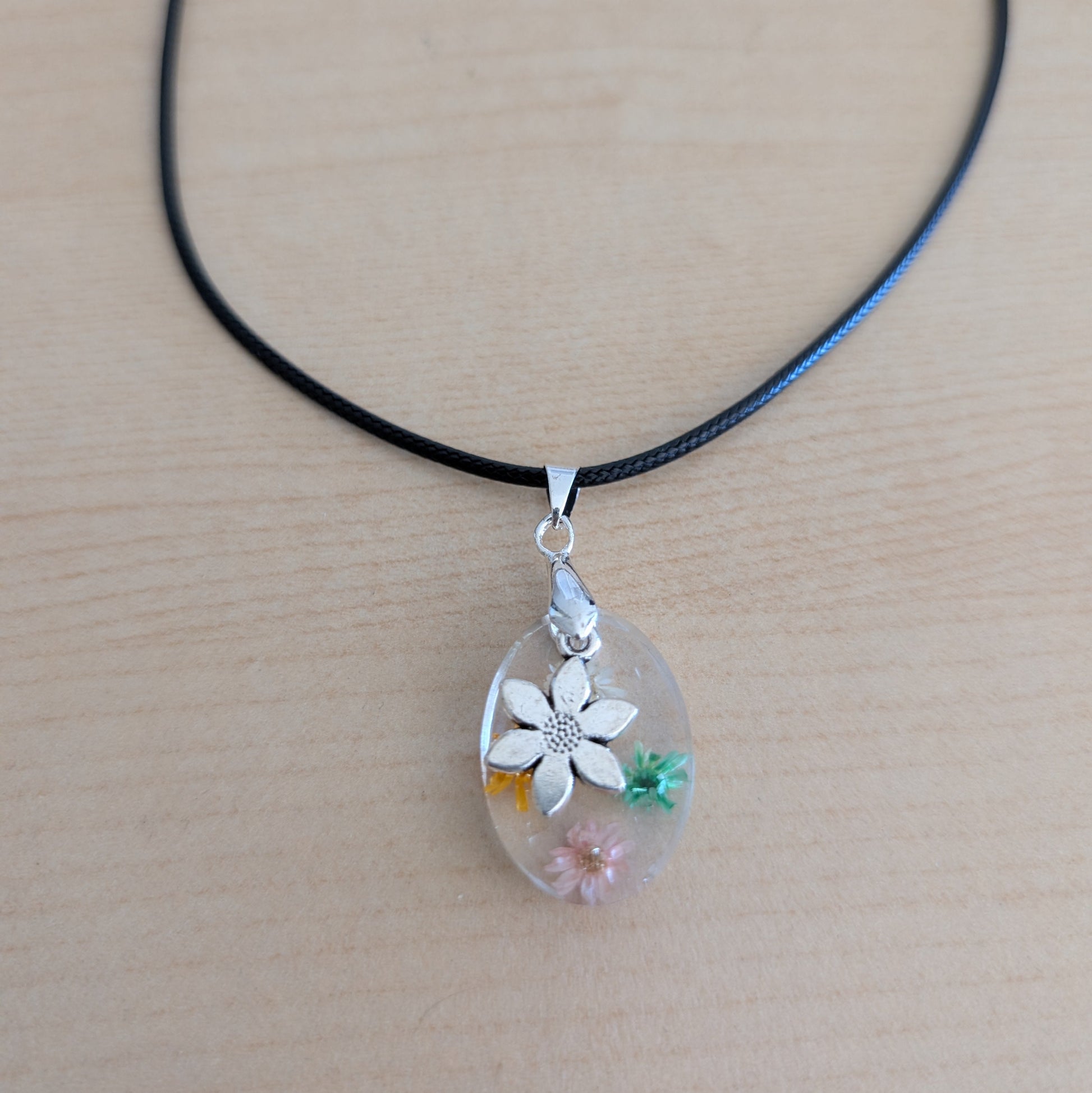 This 17.5-inch necklace showcases a stunning oval resin pendant filled with delicate flowers, complemented by a silver flower charm. Strung on a black leather cord with a secure clasp, it offers a unique blend of natural elegance and modern style, perfect for enhancing any outfit with a touch of floral beauty.