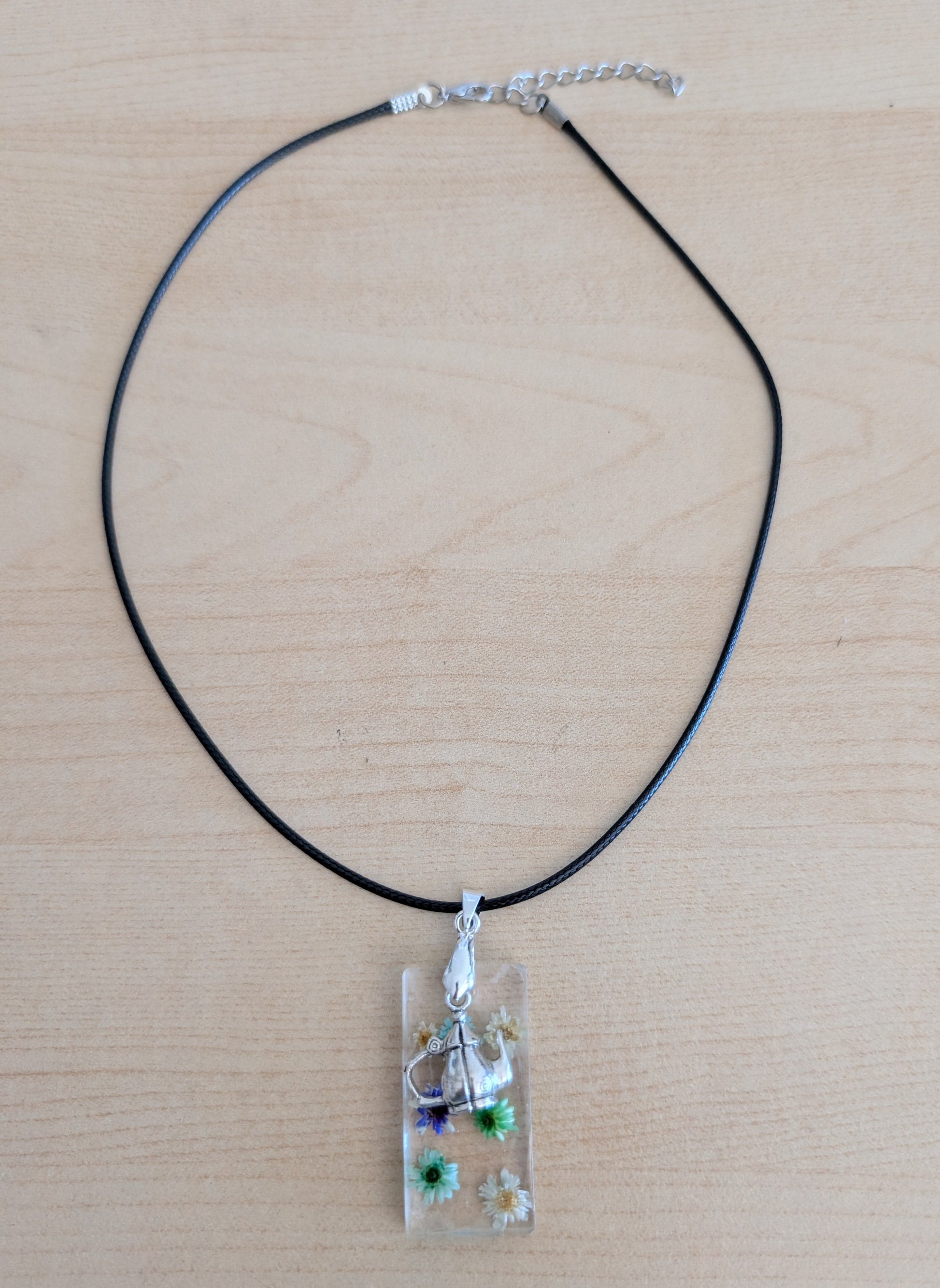 This 17.5-inch necklace features a unique rectangular resin pendant embedded with delicate flowers, paired with a charming silver kettle accent. Suspended on a sleek black leather cord with a secure clasp, it offers a stylish blend of nature-inspired beauty and modern design, perfect for adding a distinctive touch to any outfit.