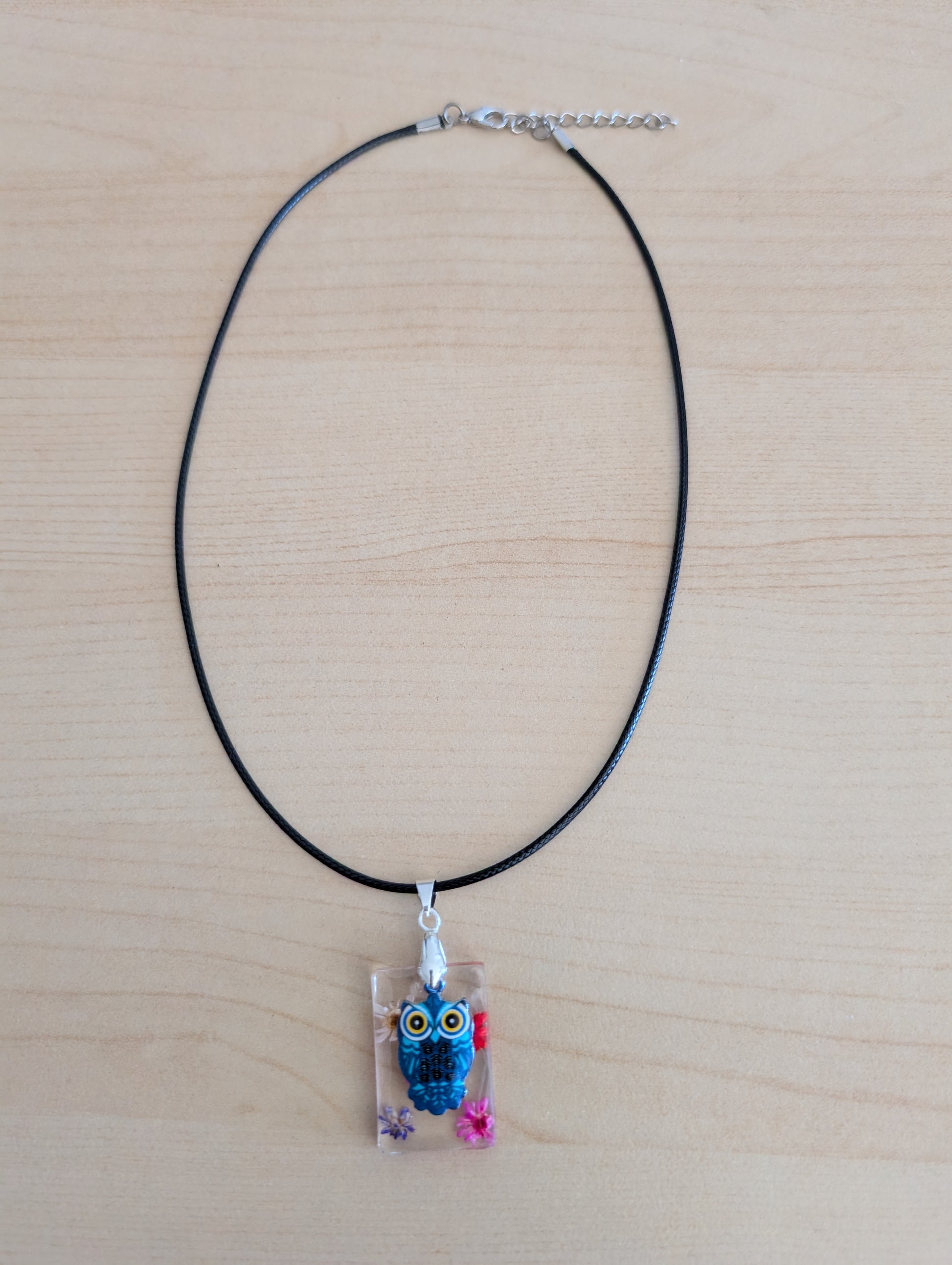 This 17.5-inch necklace features a unique rectangular resin pendant embedded with delicate flowers, paired with a charming silver owl accent. Suspended on a sleek black leather cord with a secure clasp, it combines natural beauty and subtle elegance, making it a perfect accessory to enhance any outfit.