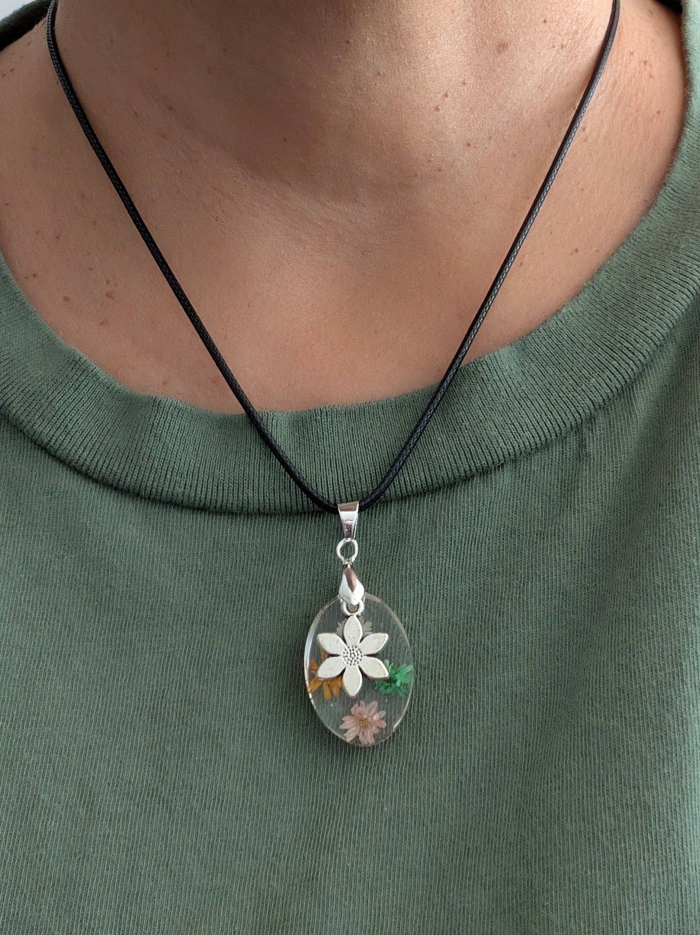This 17.5-inch necklace showcases a stunning oval resin pendant filled with delicate flowers, complemented by a silver flower charm. Strung on a black leather cord with a secure clasp, it offers a unique blend of natural elegance and modern style, perfect for enhancing any outfit with a touch of floral beauty.