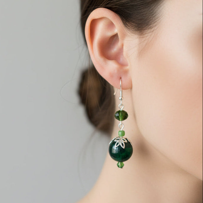 A close-up of a model's ear wearing a dangling earring with dark green round beads, small green spacer beads, and a silver-toned floral accent.