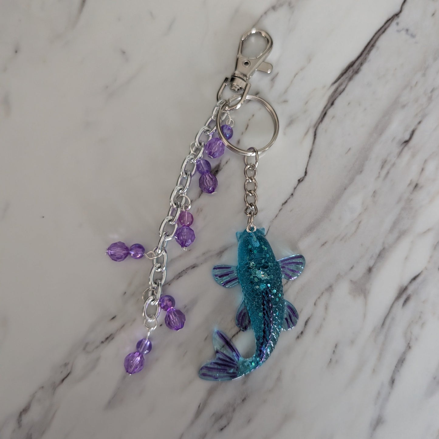 Keychain with blue glittery fish charm and purple beads on a marble surface