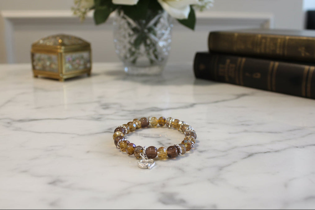 Express your heart with this warm and radiant elastic bracelet that combines rich brown and gold tones with timeless sentiment. Featuring 8mm brown and gold beads interspersed with sparkling crystal and glass accents, silver spacers, and a delicate silver heart charm, this piece speaks to both style and emotion. The elastic band stretches effortlessly to fit any wrist, requiring no clasps or adjustments—just slip it on and go. Weighing a comfortable 0.60 oz, it's substantial enough to feel quality yet light