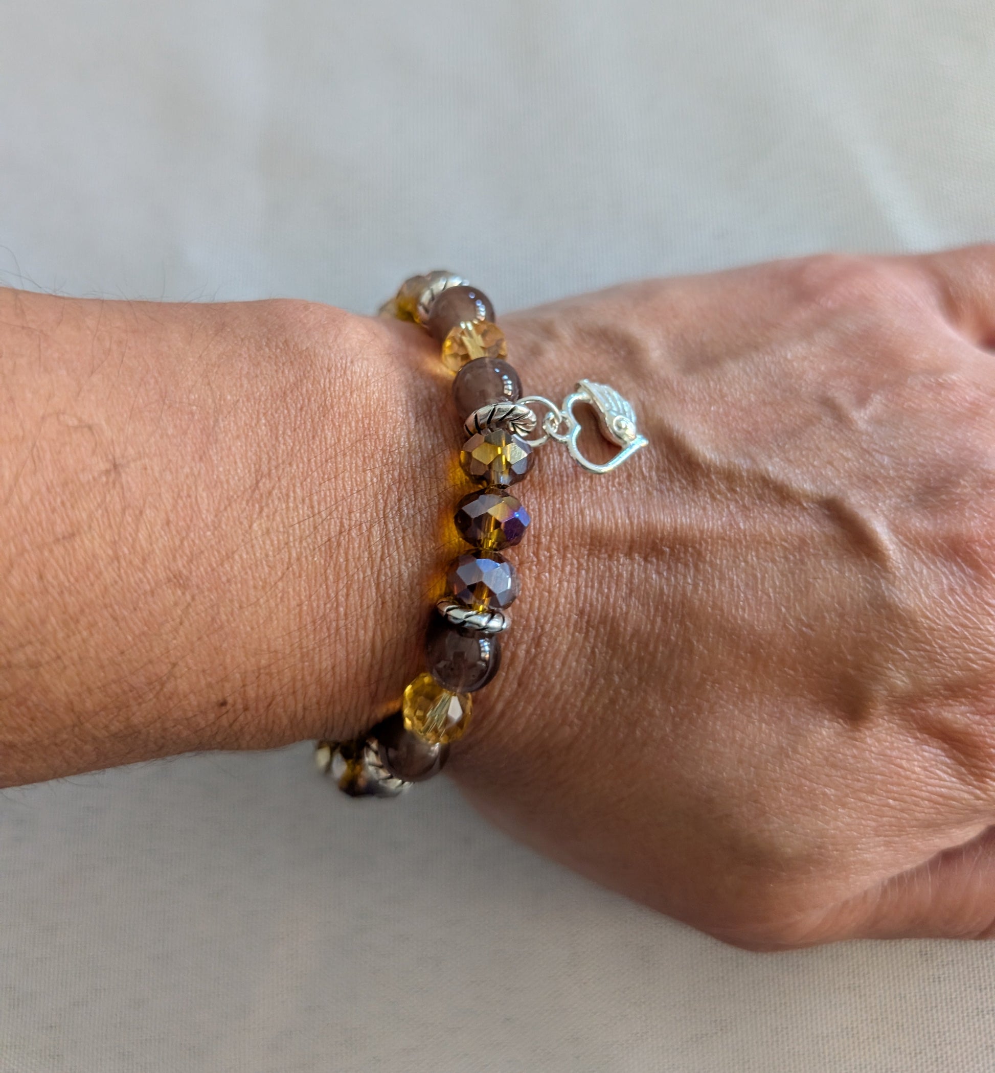 Express your heart with this warm and radiant elastic bracelet that combines rich brown and gold tones with timeless sentiment. Featuring 8mm brown and gold beads interspersed with sparkling crystal and glass accents, silver spacers, and a delicate silver heart charm, this piece speaks to both style and emotion. The elastic band stretches effortlessly to fit any wrist, requiring no clasps or adjustments—just slip it on and go. Weighing a comfortable 0.60 oz, it's substantial enough to feel quality yet light