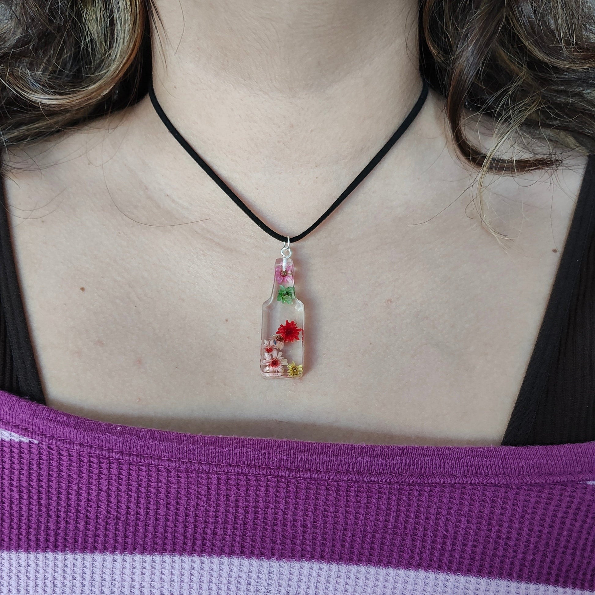 Wear our stunning 15.5 inch black suede cord necklace with a unique resin bottle pendant adorned with delicate flowers. Elevate your style and add a touch of whimsy to any outfit. Perfect for any occasion, this necklace is a must-have for any fashion-forward individual.