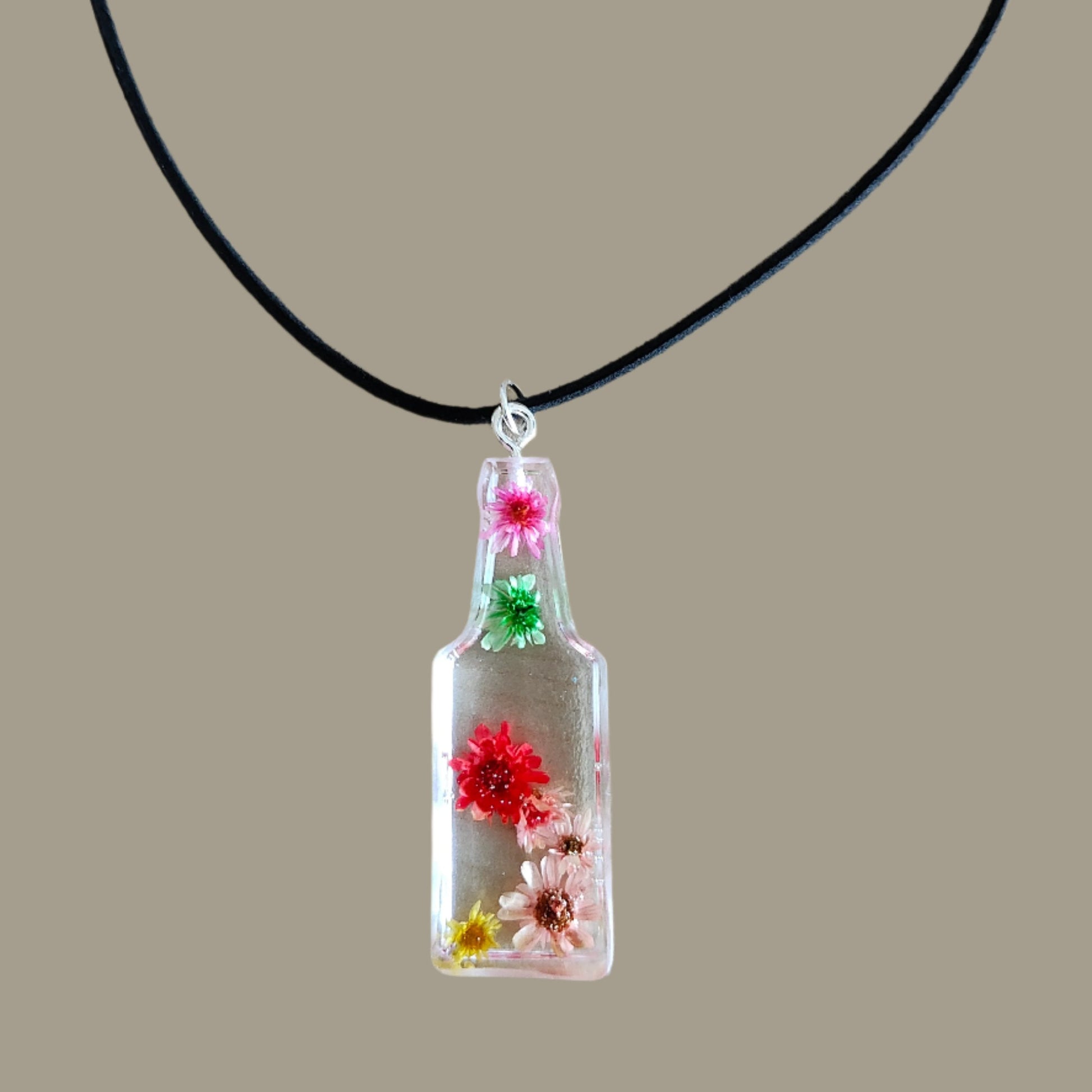 Wear our stunning 15.5 inch black suede cord necklace with a unique resin bottle pendant adorned with delicate flowers. Elevate your style and add a touch of whimsy to any outfit. Perfect for any occasion, this necklace is a must-have for any fashion-forward individual.