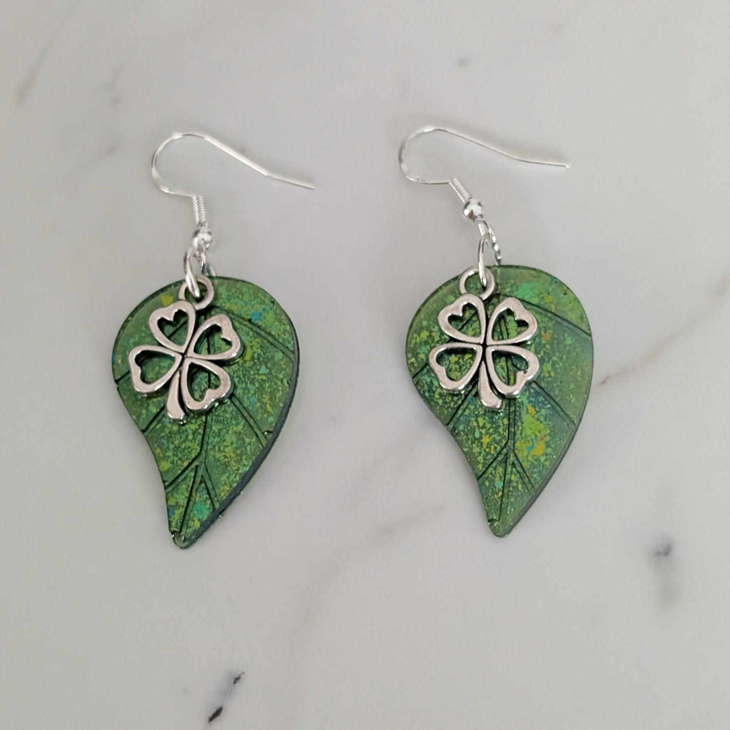 Pair of green leaf-shaped earrings with silver four-leaf clover charms on a marble surface.