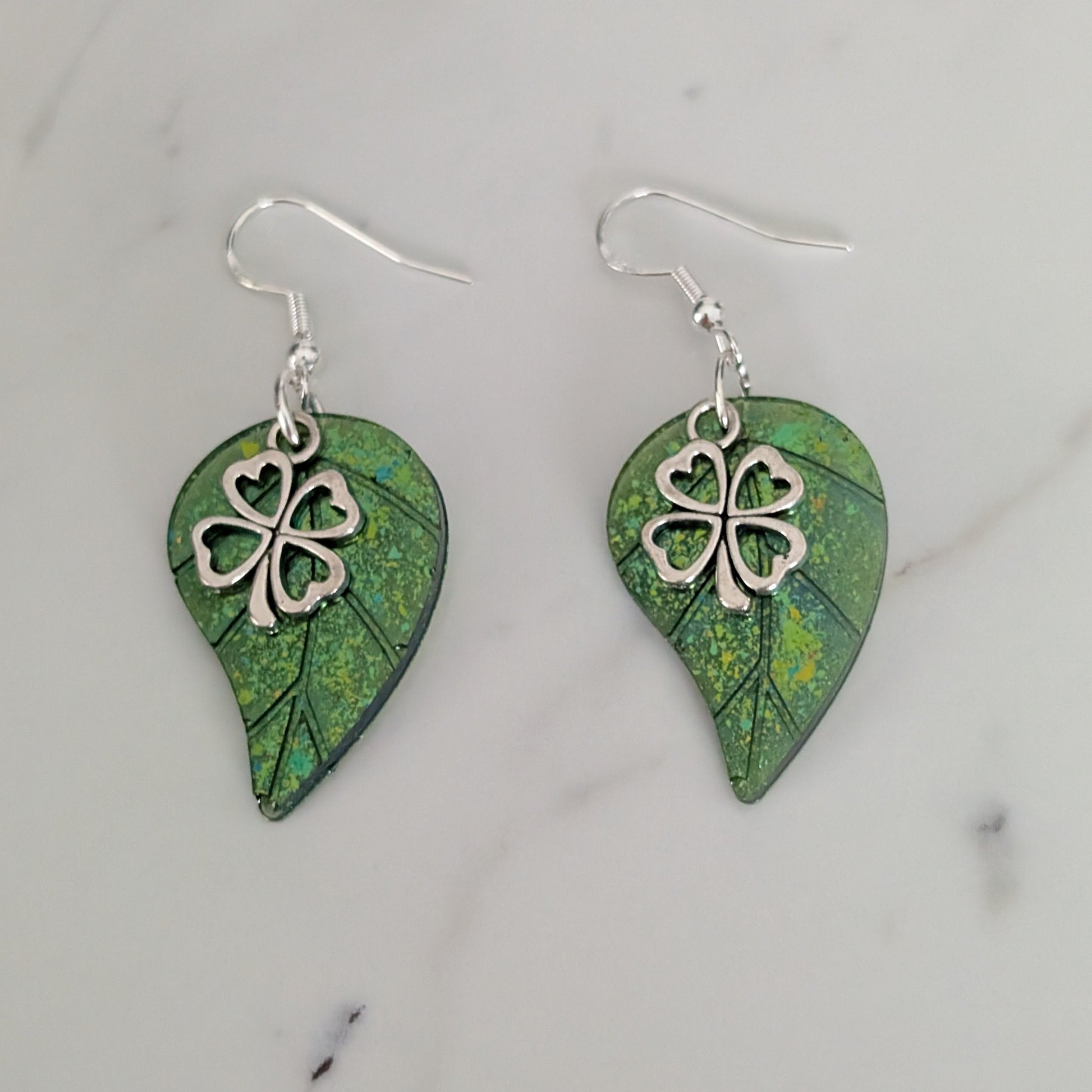 Pair of green leaf-shaped earrings with silver four-leaf clover charms on a marble surface.