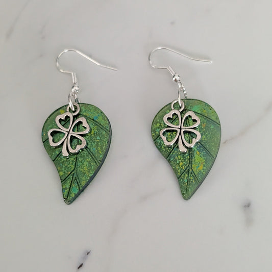 Pair of green leaf-shaped earrings with silver four-leaf clover charms on a marble surface.