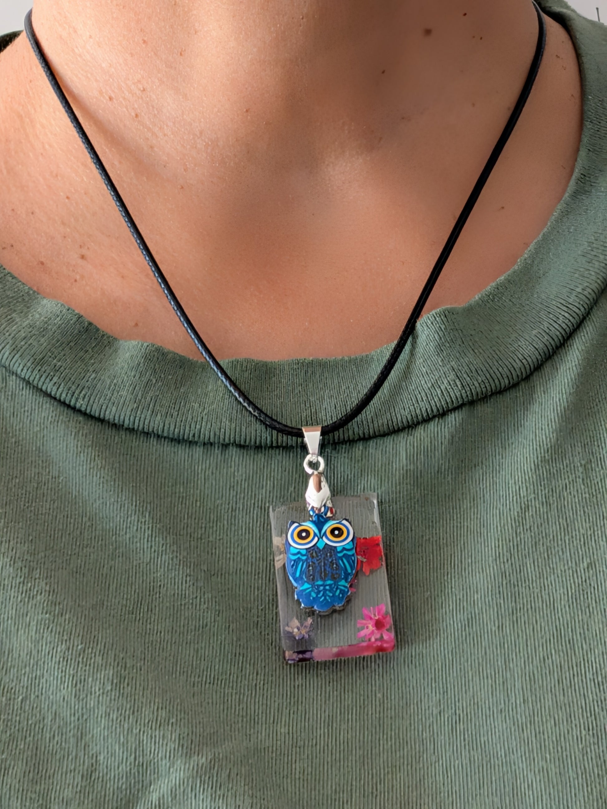 This 17.5-inch necklace features a unique rectangular resin pendant embedded with delicate flowers, paired with a charming silver owl accent. Suspended on a sleek black leather cord with a secure clasp, it combines natural beauty and subtle elegance, making it a perfect accessory to enhance any outfit.