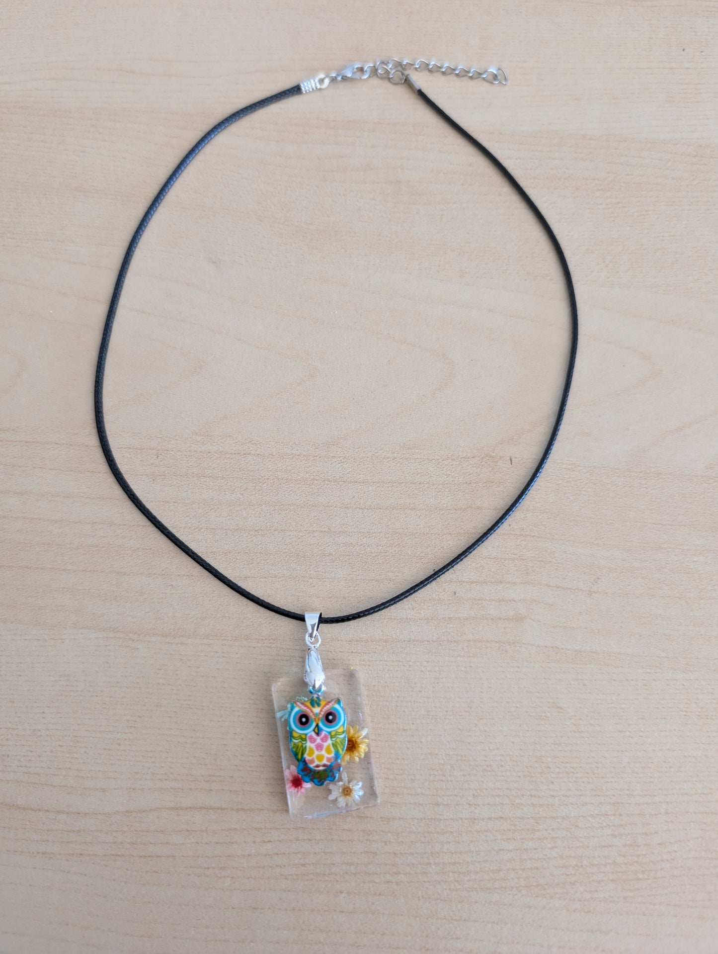 This 17.5-inch necklace features a unique rectangular resin pendant embedded with delicate flowers, paired with a charming silver owl accent. Suspended on a sleek black leather cord with a secure clasp, it combines natural beauty and subtle elegance, making it a perfect accessory to enhance any outfit.