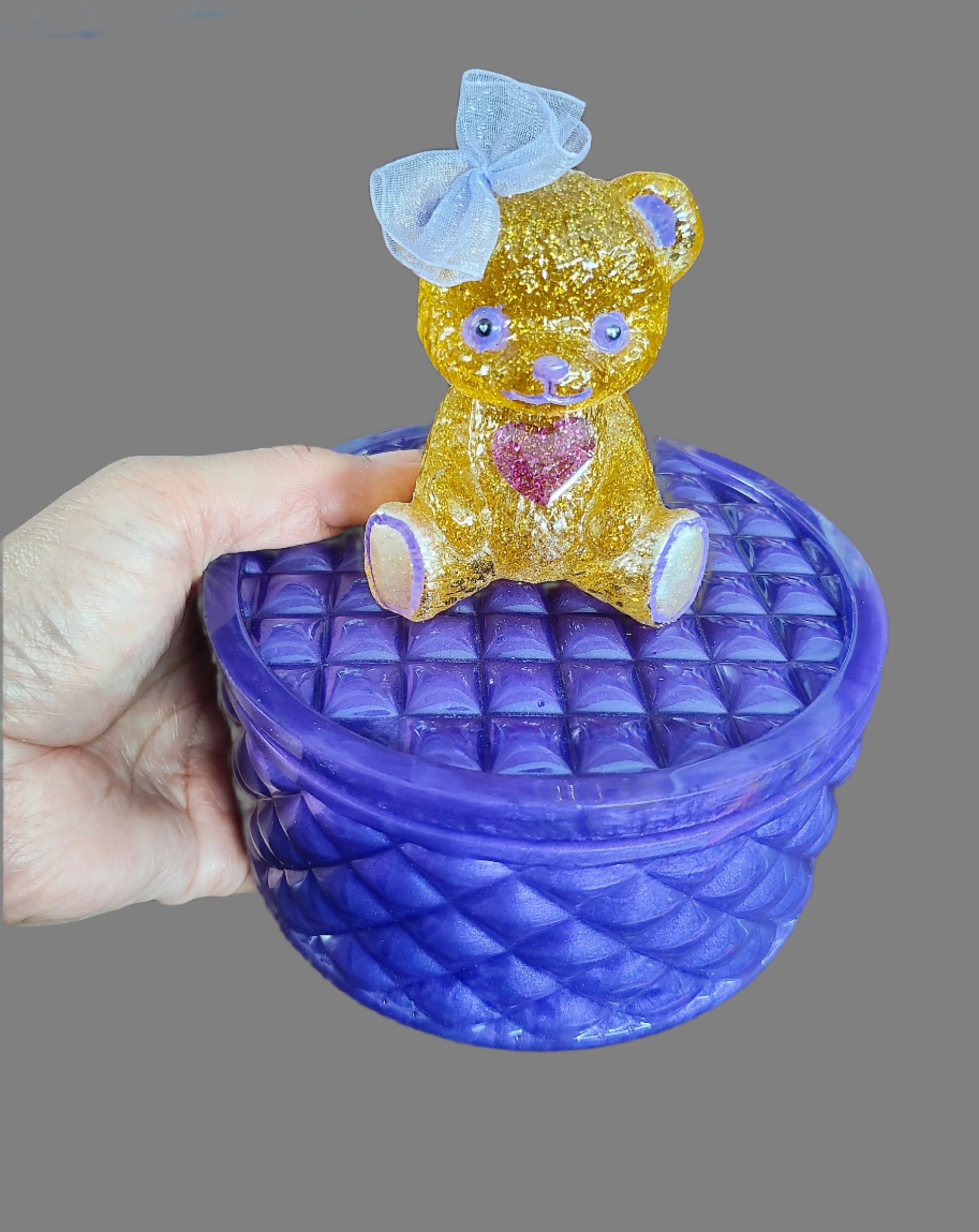 This adorable hand-crafted resin trinket box brings a touch of whimsy to your space! Featuring a charming teddy bear perched on top, it's the perfect home for jewelry, keepsakes, or small treasures. Measuring 5" x 5" x 5" with a 2" deep dish, this compact beauty combines functionality with personality. Crafted from smooth, durable epoxy resin that feels like glass, it's built to last while adding a playful accent to any dresser or shelf. At just 13 oz, it's lightweight enough to move around but sturdy enoug
