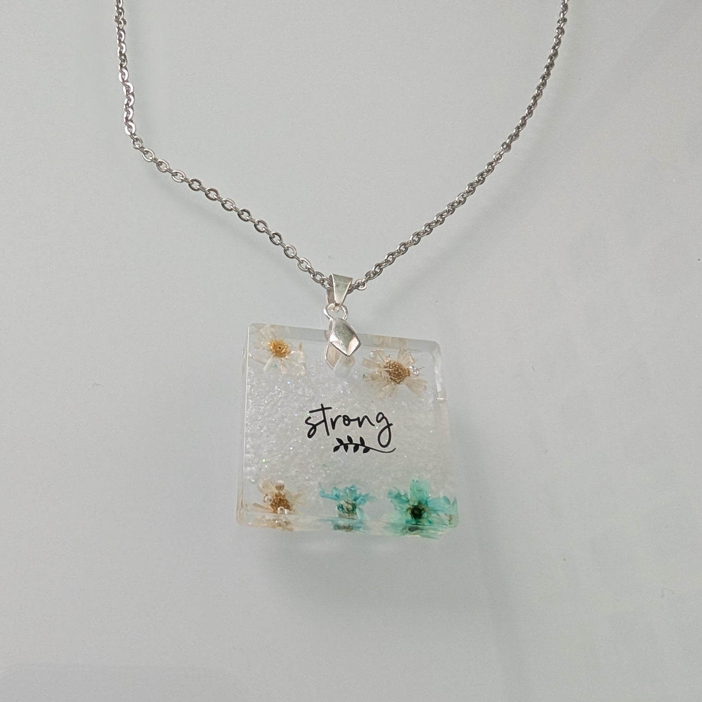 This stainless steel cable link chain necklace features a beautiful square pendant with real flowers embedded in resin. The necklace is 21 inches long, with an additional 2 inch extension chain. Make a statement with this one-of-a-kind piece!

Necklace - 21 inches stainless steel cable link chain with a resin with flowers square pendant