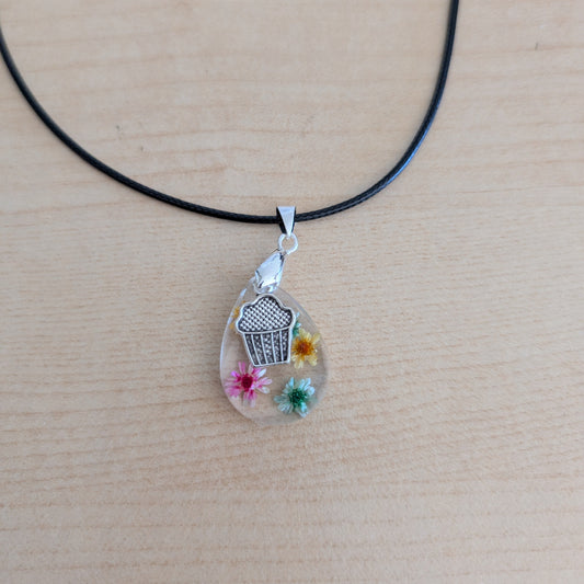 This charming 17.5-inch necklace showcases a resin teardrop filled with delicate flowers, paired with a playful silver cupcake charm. Set on a black leather cord with a secure clasp, it combines whimsical detail with stylish simplicity, making it a unique accessory to brighten any look.