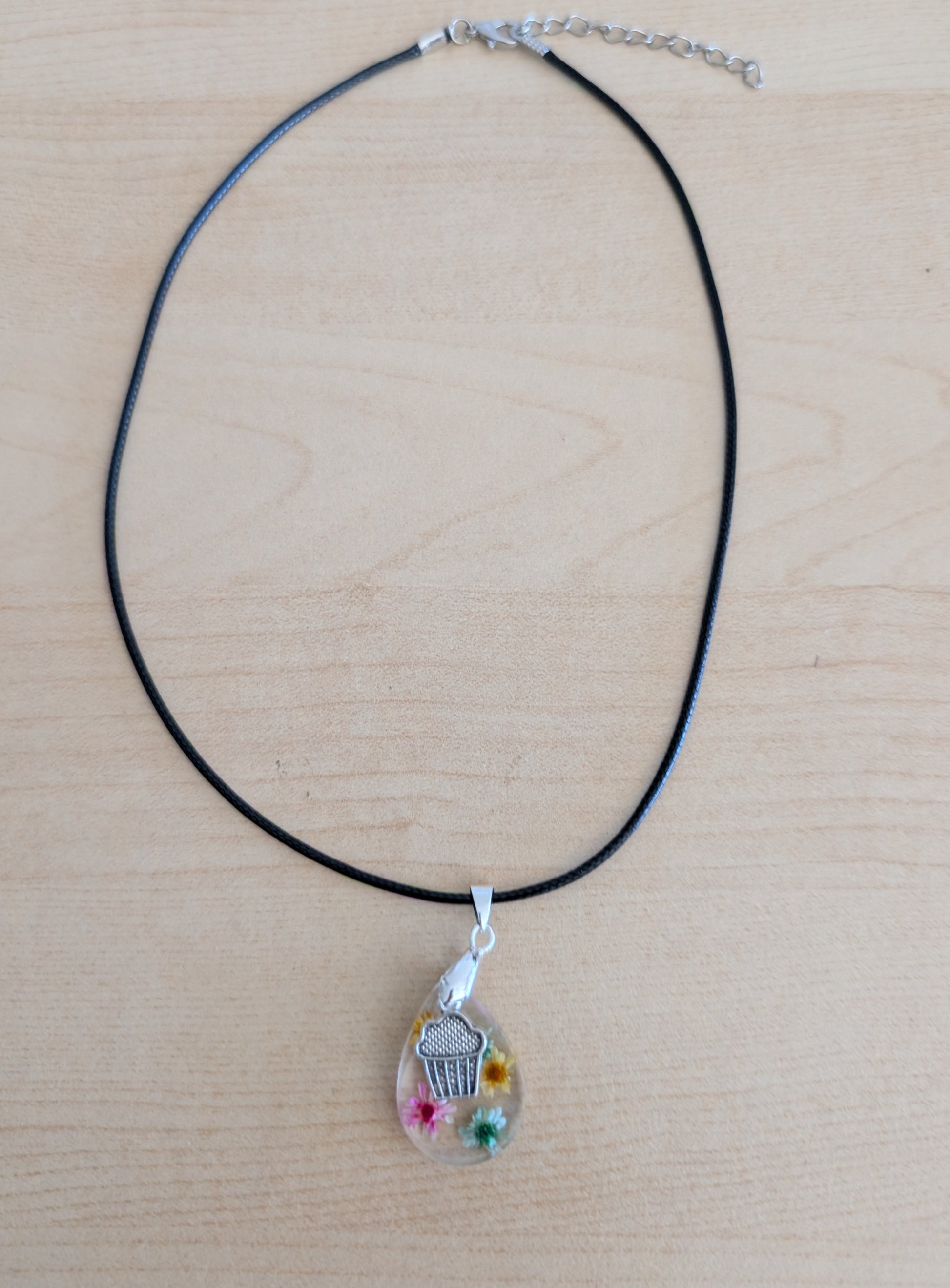 This charming 17.5-inch necklace showcases a resin teardrop filled with delicate flowers, paired with a playful silver cupcake charm. Set on a black leather cord with a secure clasp, it combines whimsical detail with stylish simplicity, making it a unique accessory to brighten any look.