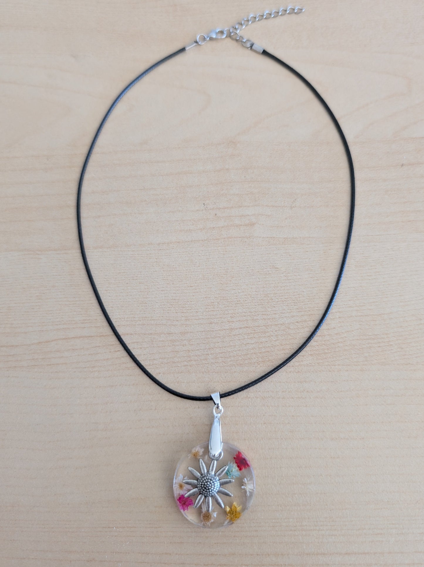 This elegant 17.5-inch necklace features a delicate resin circle embedded with vibrant flowers, complemented by a silver flower charm. Suspended on a sleek black leather cord with a secure clasp, it offers a perfect blend of natural beauty and modern style. Ideal for adding a subtle yet captivating touch to any outfit.