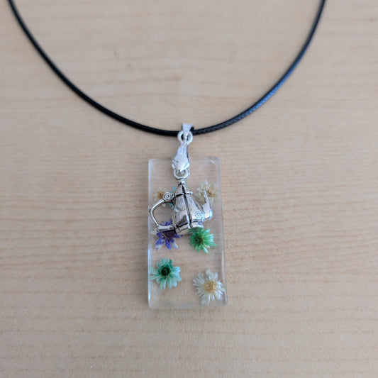 This 17.5-inch necklace features a unique rectangular resin pendant embedded with delicate flowers, paired with a charming silver kettle accent. Suspended on a sleek black leather cord with a secure clasp, it offers a stylish blend of nature-inspired beauty and modern design, perfect for adding a distinctive touch to any outfit.