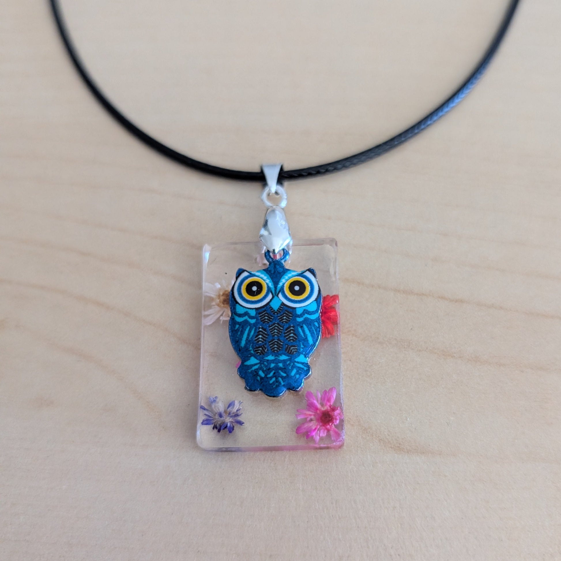 This 17.5-inch necklace features a unique rectangular resin pendant embedded with delicate flowers, paired with a charming silver owl accent. Suspended on a sleek black leather cord with a secure clasp, it combines natural beauty and subtle elegance, making it a perfect accessory to enhance any outfit.